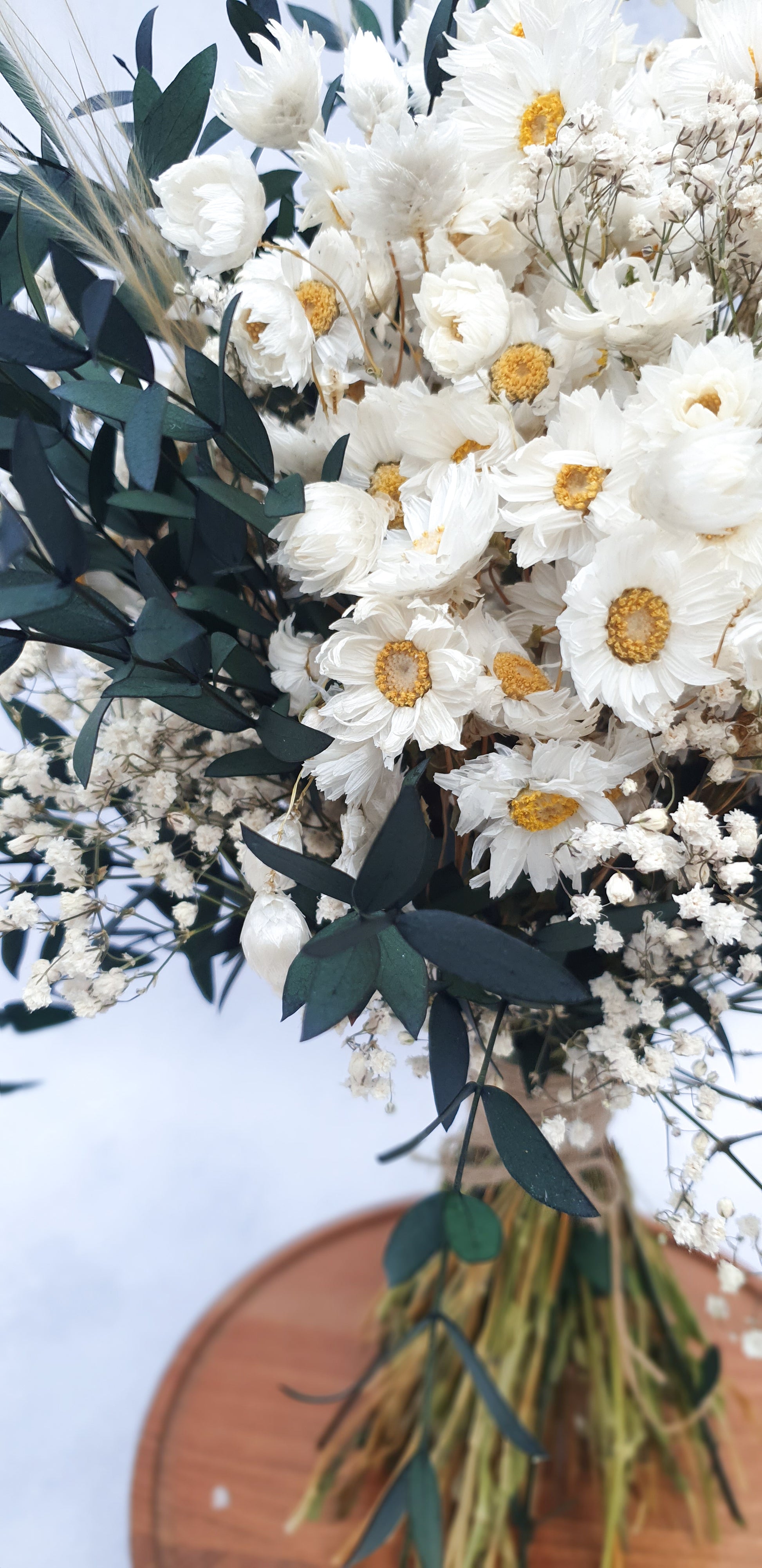 "Snowdrop Glade" Minimalist Large Handtie Bouquet - Lytton Rose Botanical. A detailed close-up of a dried flower bouquet. Delicate white daisies with yellow centers take center stage, surrounded by lush green eucalyptus leaves and wispy baby's breath. The bouquet is elegantly tied with twine and rests on a rustic wooden board